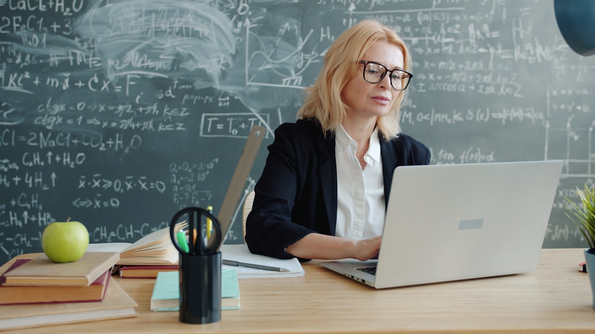 Lehrerin arbeitet am Laptop vor einer Tafel mit Formeln – Symbolbild für den Workshop „KI im Unterricht